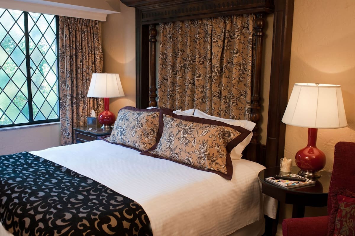 A hotel room with a bed, two bedside lamps, a window with curtains, and a view of greenery outside.