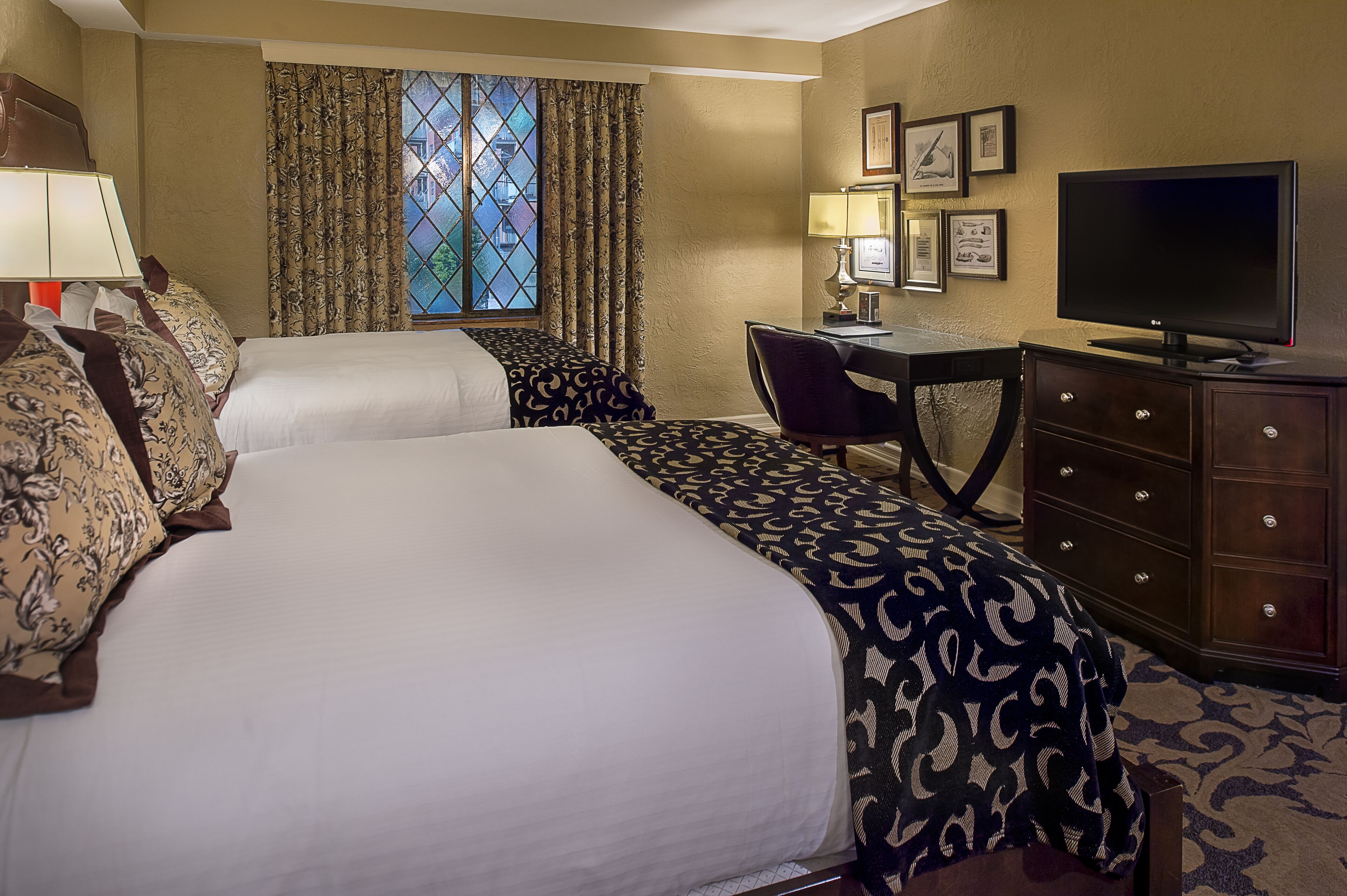 A hotel room with two beds, a desk, a television, and a window with curtains.