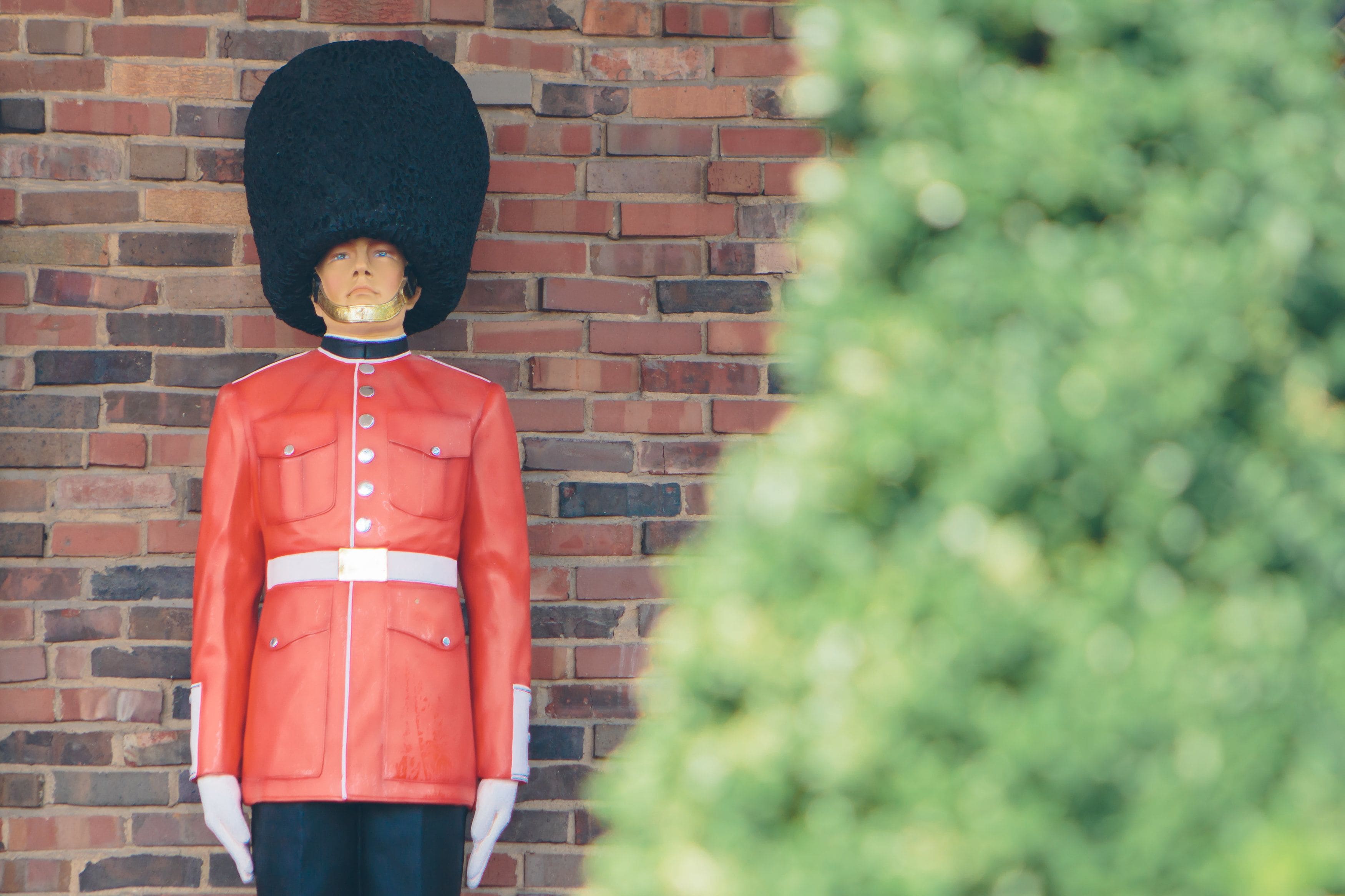A statue of a guard in a red uniform with a tall black hat, standing against a brick wall.