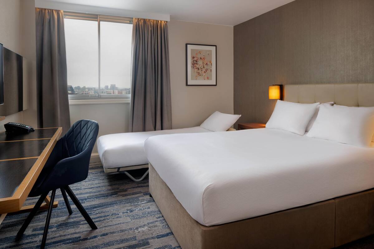 A hotel room with a bed, a desk, a chair, a lamp, and a window with curtains.