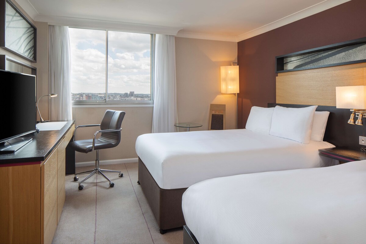 A hotel room with two beds, a desk, a chair, a TV, a lamp, and a view of the city.