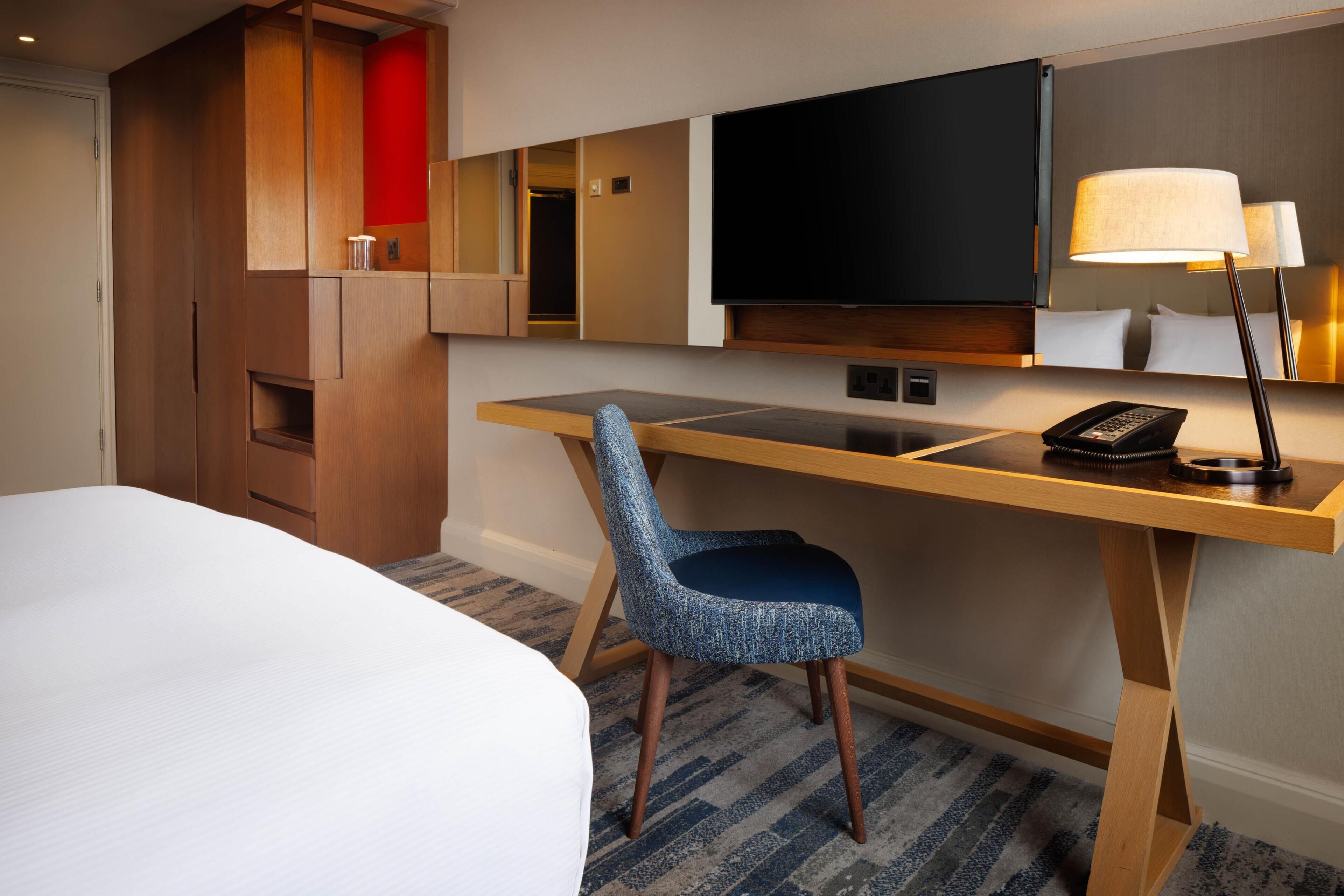 A hotel room with a bed, a desk, a chair, a TV, and a lamp.