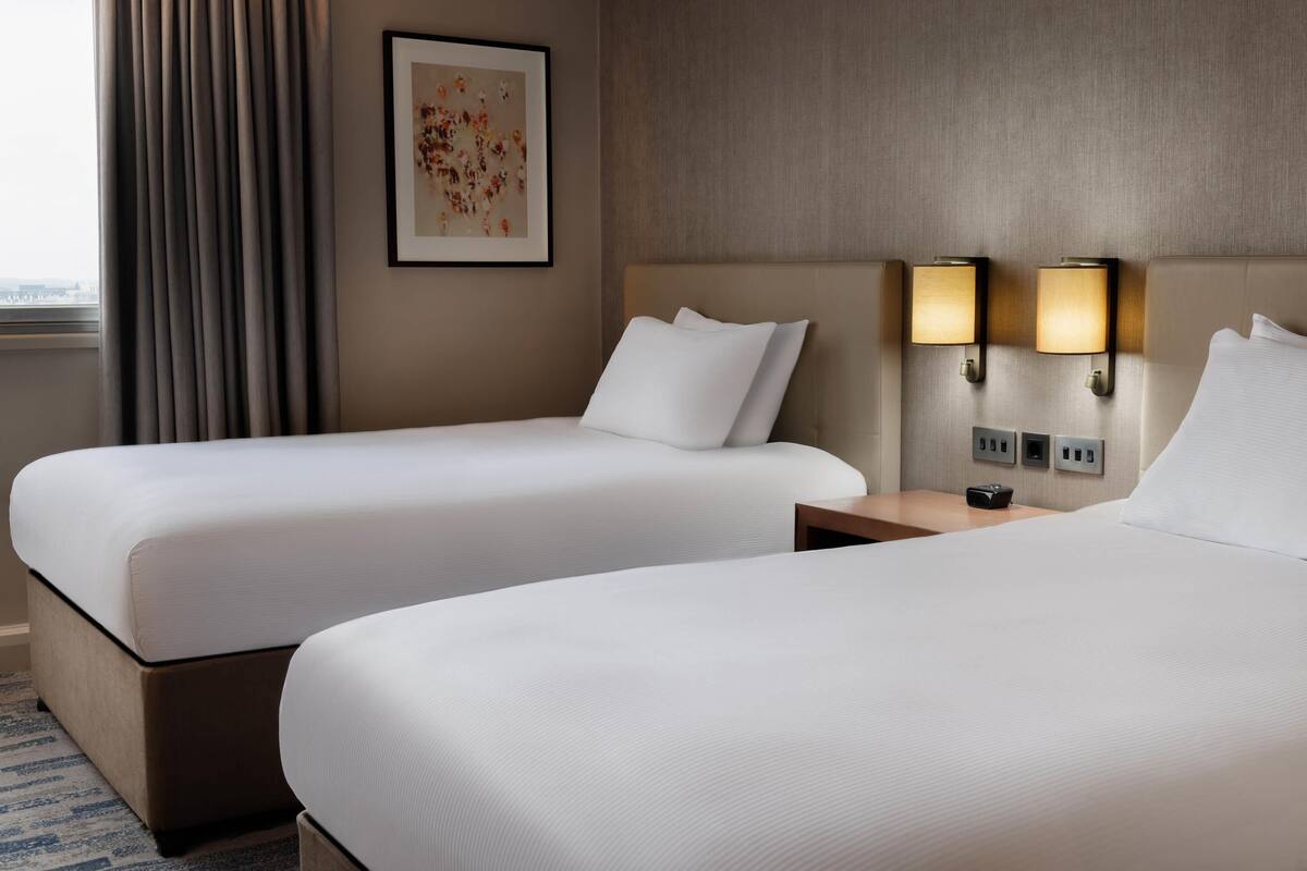 A hotel room with two beds, a framed artwork on the wall, and wall-mounted lamps.