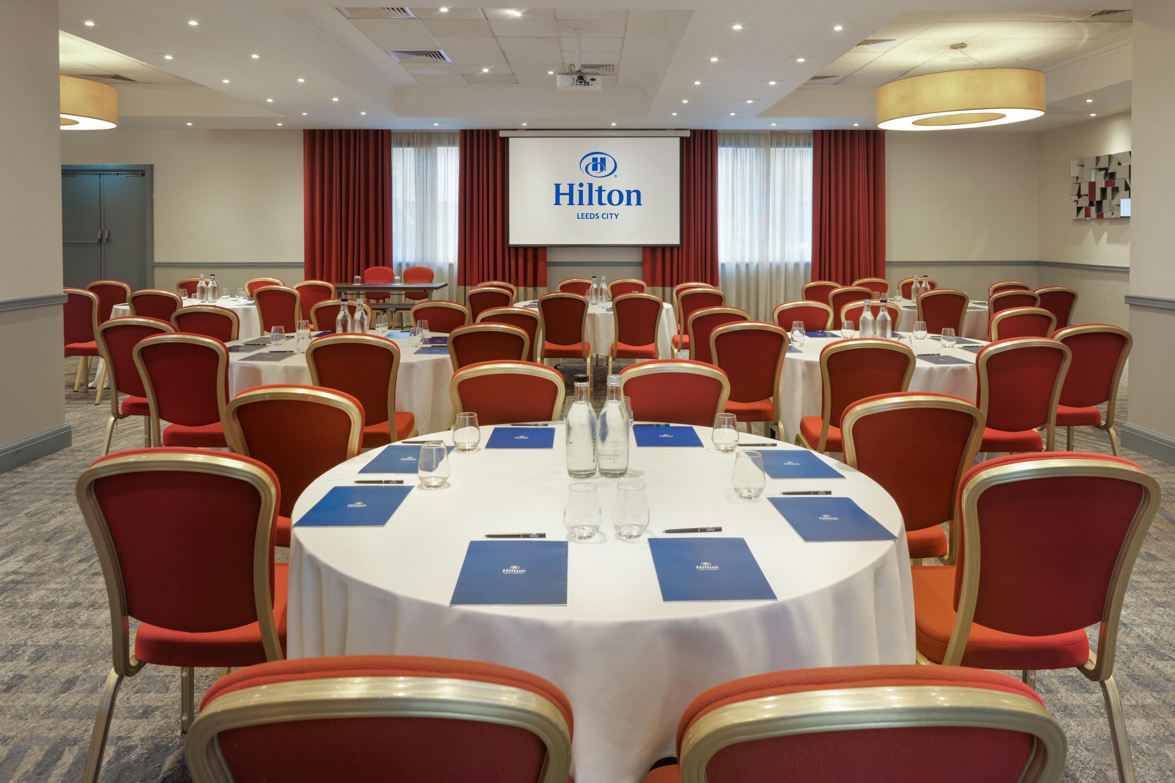 A conference room with round tables, red chairs, and a large screen displaying the Hilton logo.