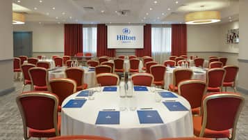 A conference room with round tables, red chairs, and a large screen displaying the Hilton logo.