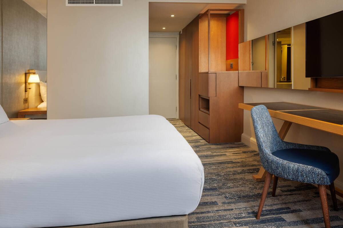 A hotel room with a large bed, a desk, a chair, a TV, and a wardrobe.