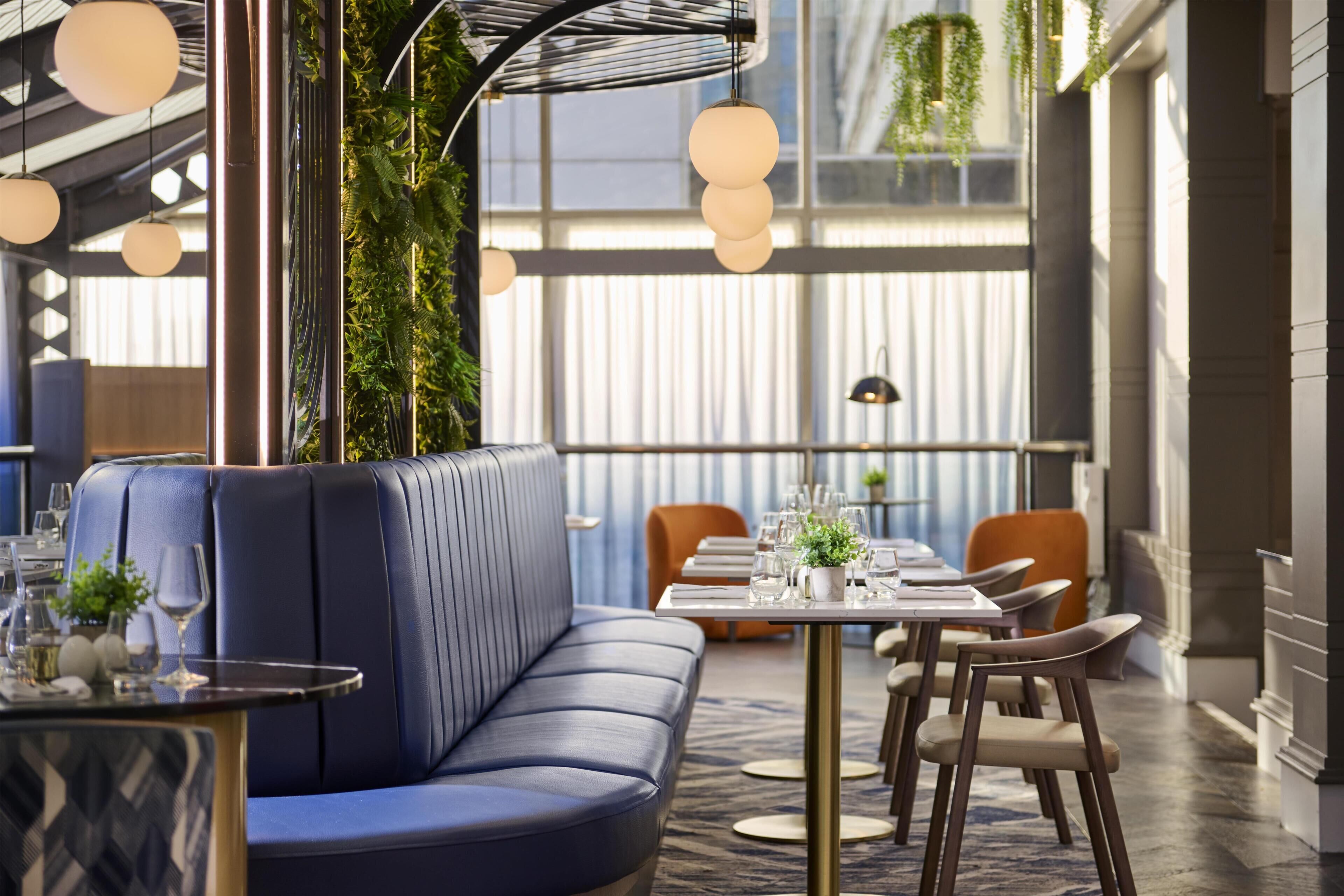 A modern restaurant with a blue leather booth, round tables, and hanging plants.