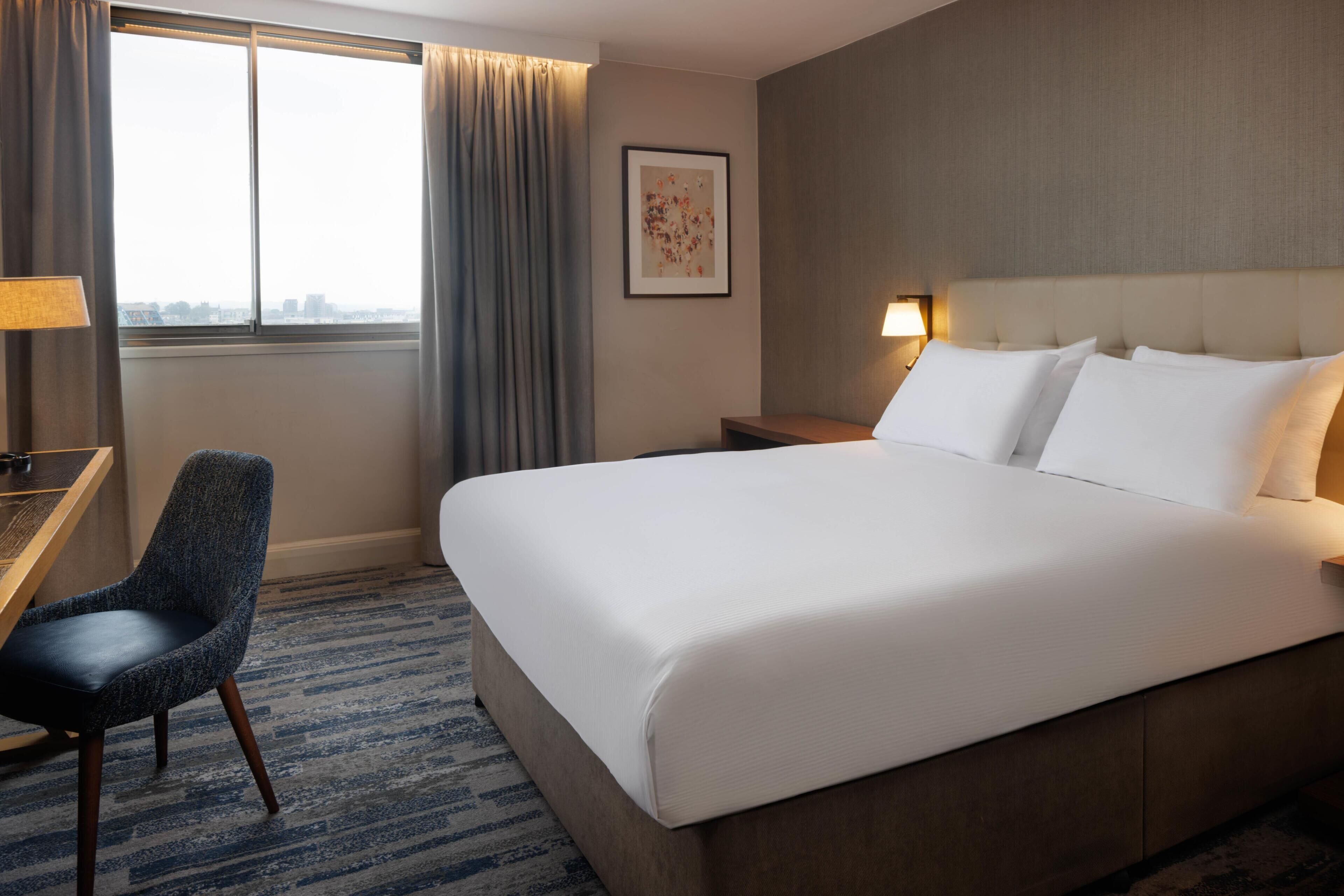A hotel room with a large bed, a desk with a chair, a window with curtains, a framed picture on the wall, and a nightstand with a lamp.