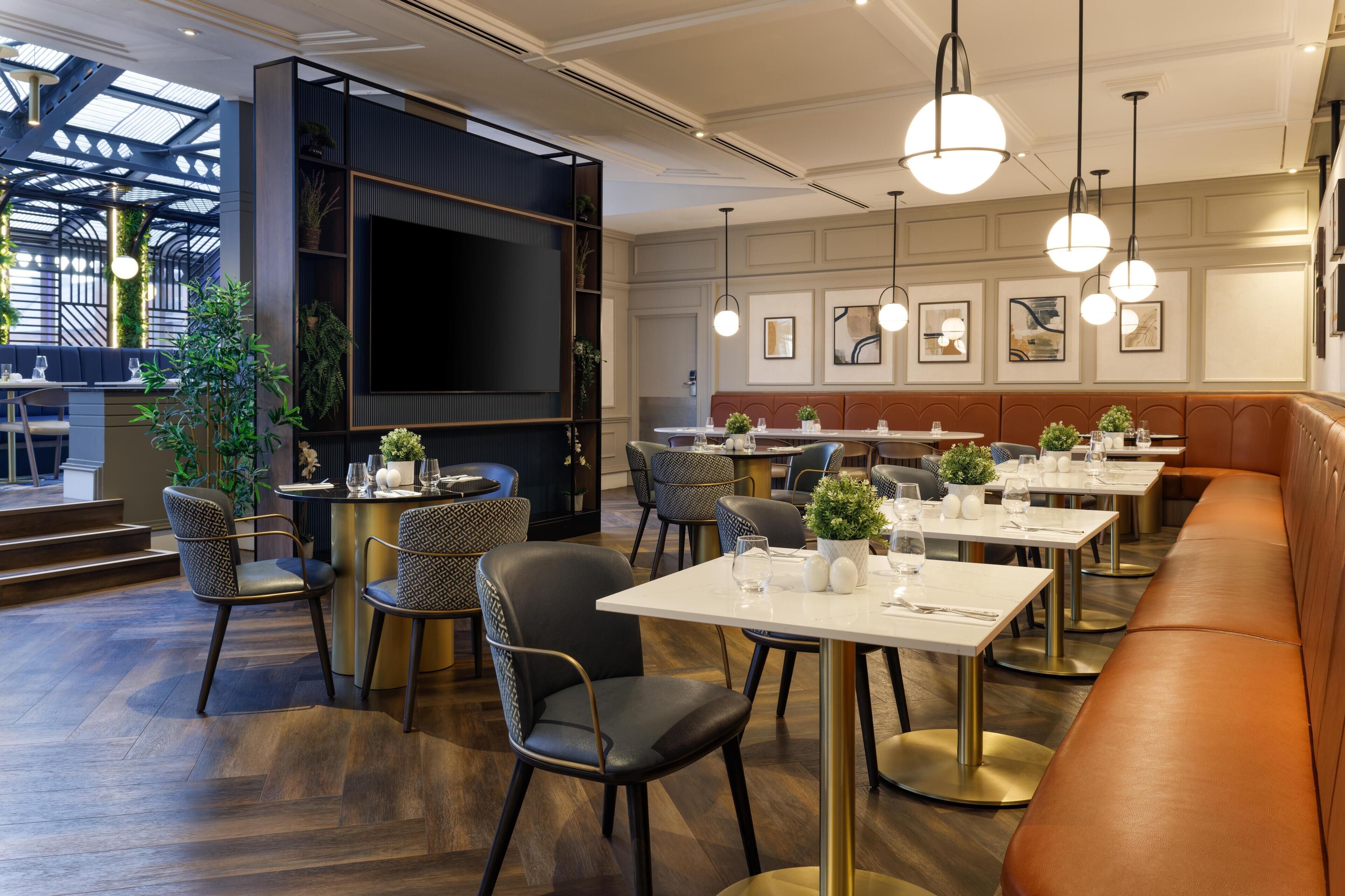 Een modern restaurant met hoge plafonds, een groot televisiescherm en diverse zitmogelijkheden, waaronder tafels en zitbankjes.