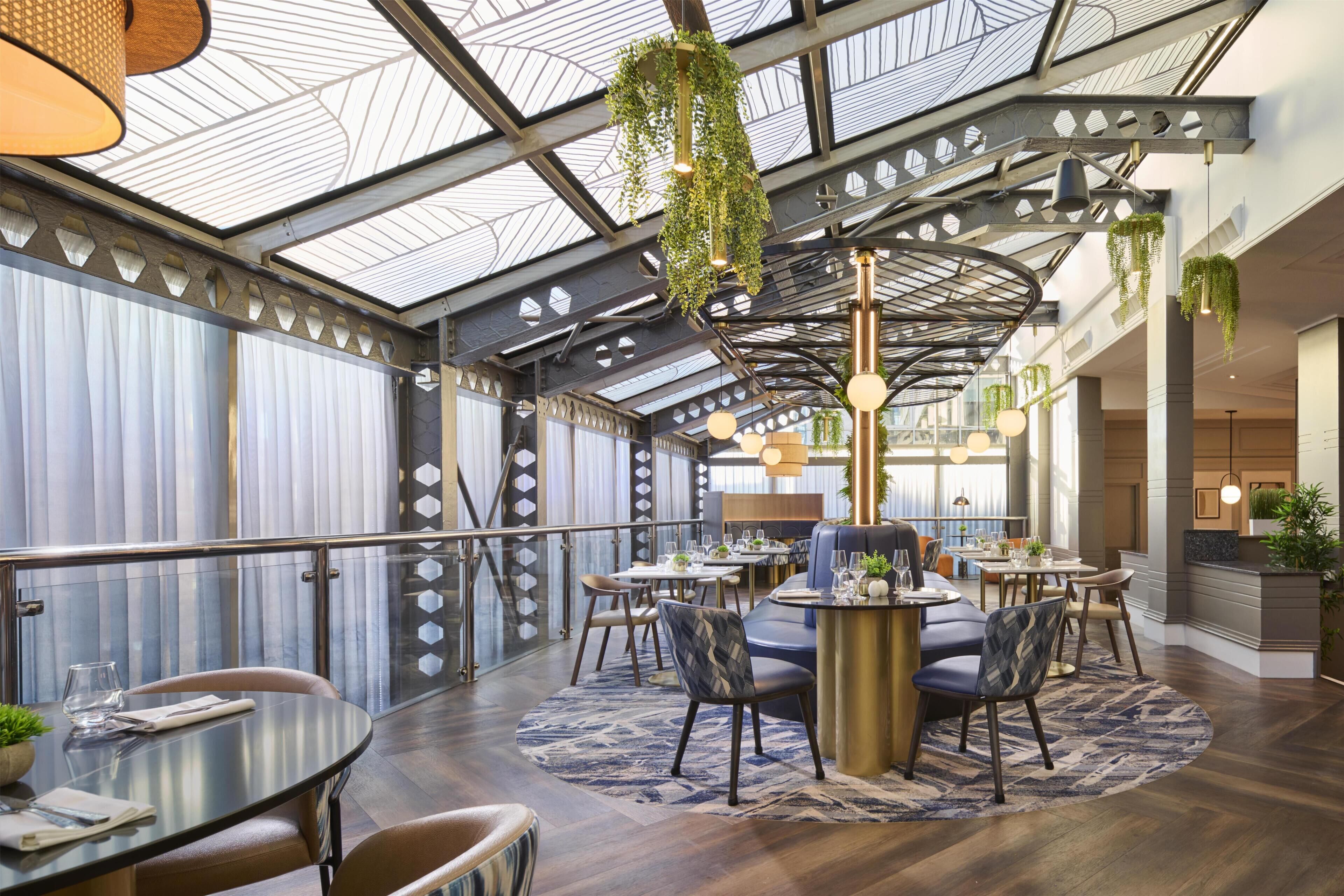 A spacious indoor area with multiple dining tables and chairs, a glass ceiling, and hanging plants.