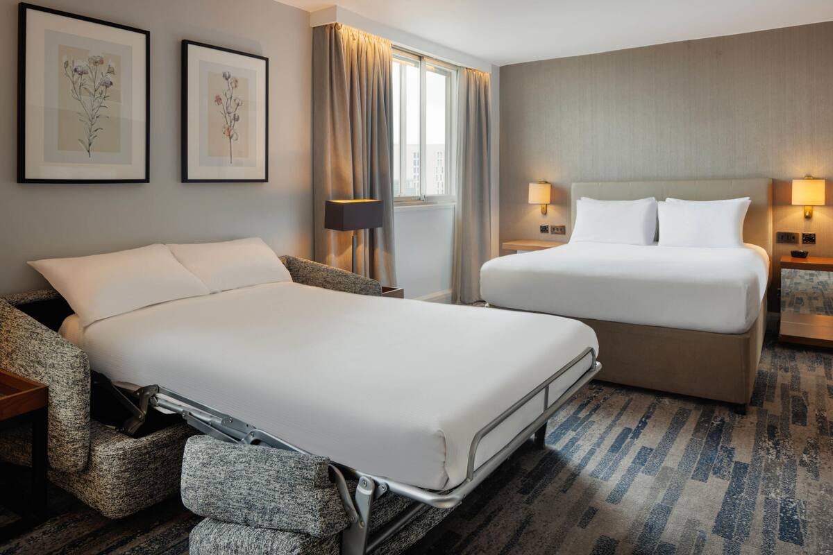 A hotel room with two beds, a sofa, a lamp, and framed artwork on the wall.