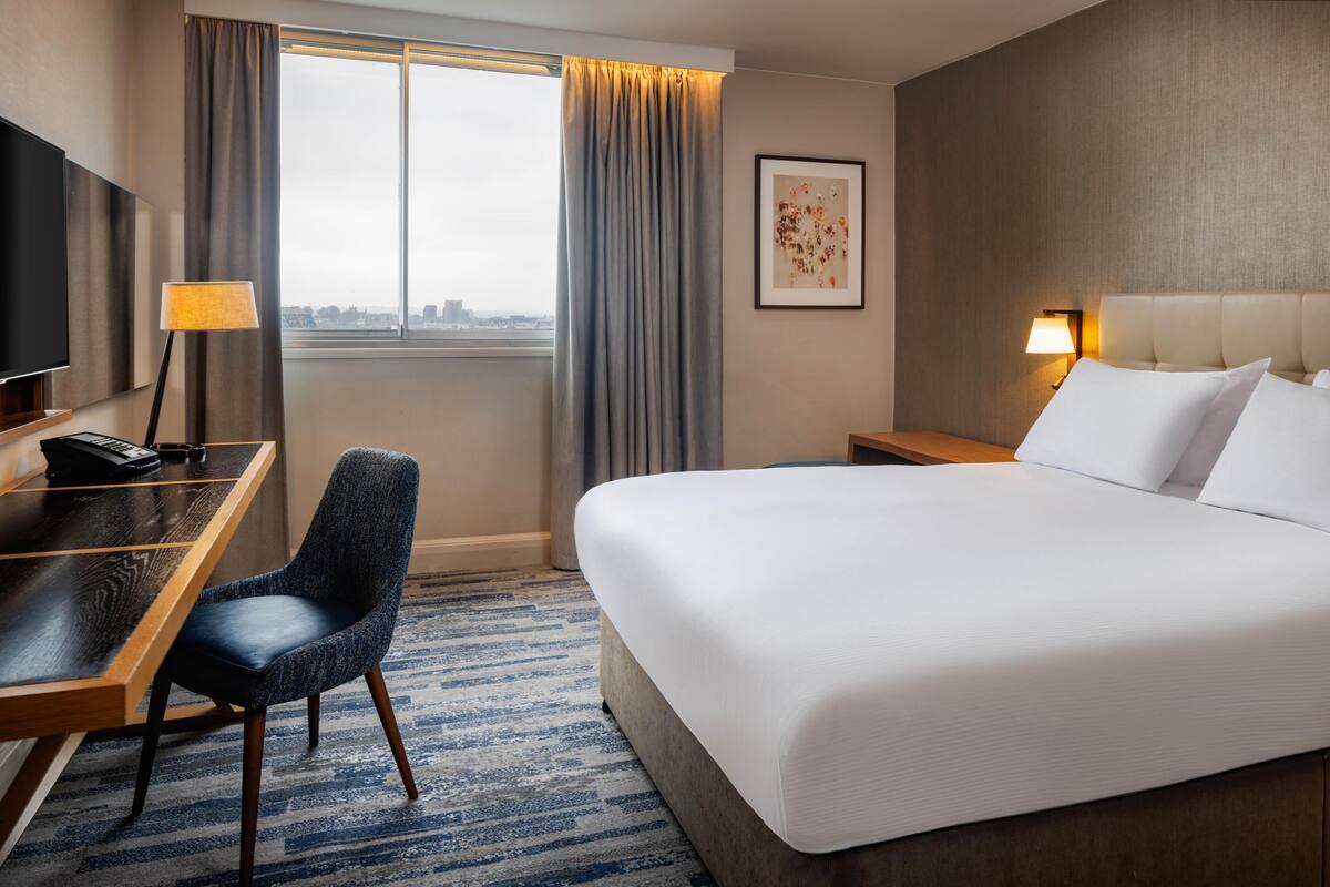 A hotel room with a bed, desk, chair, and a city view.