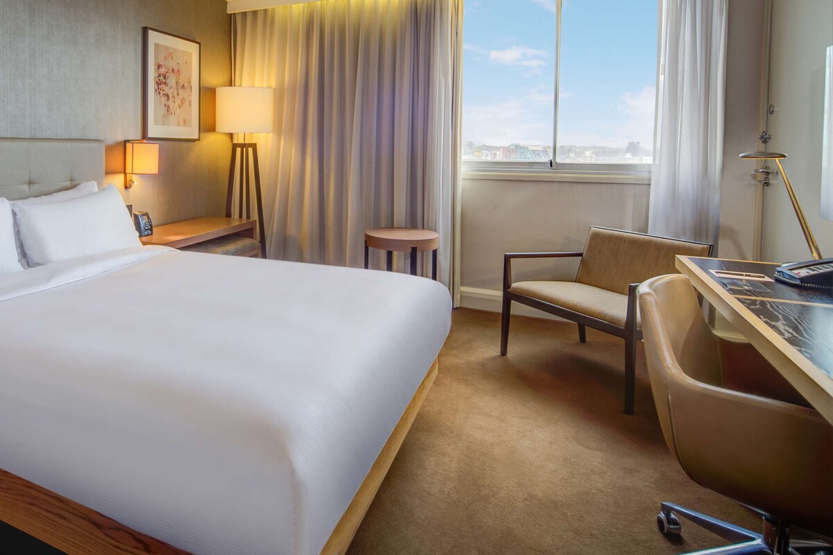A hotel room with a large bed, a desk with a chair, a small stool, a lamp, and a window with a view.