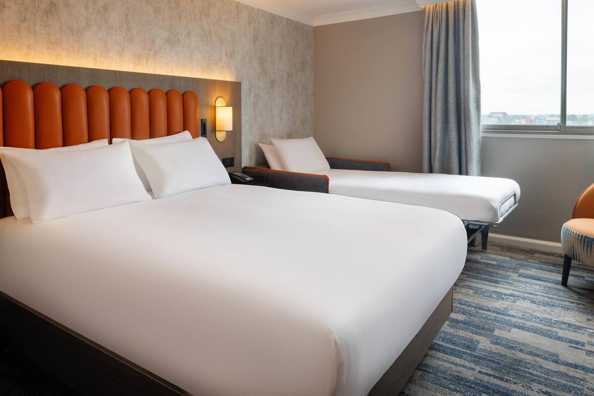 A hotel room with two beds, a headboard, a nightstand, a chair, and a window with curtains.