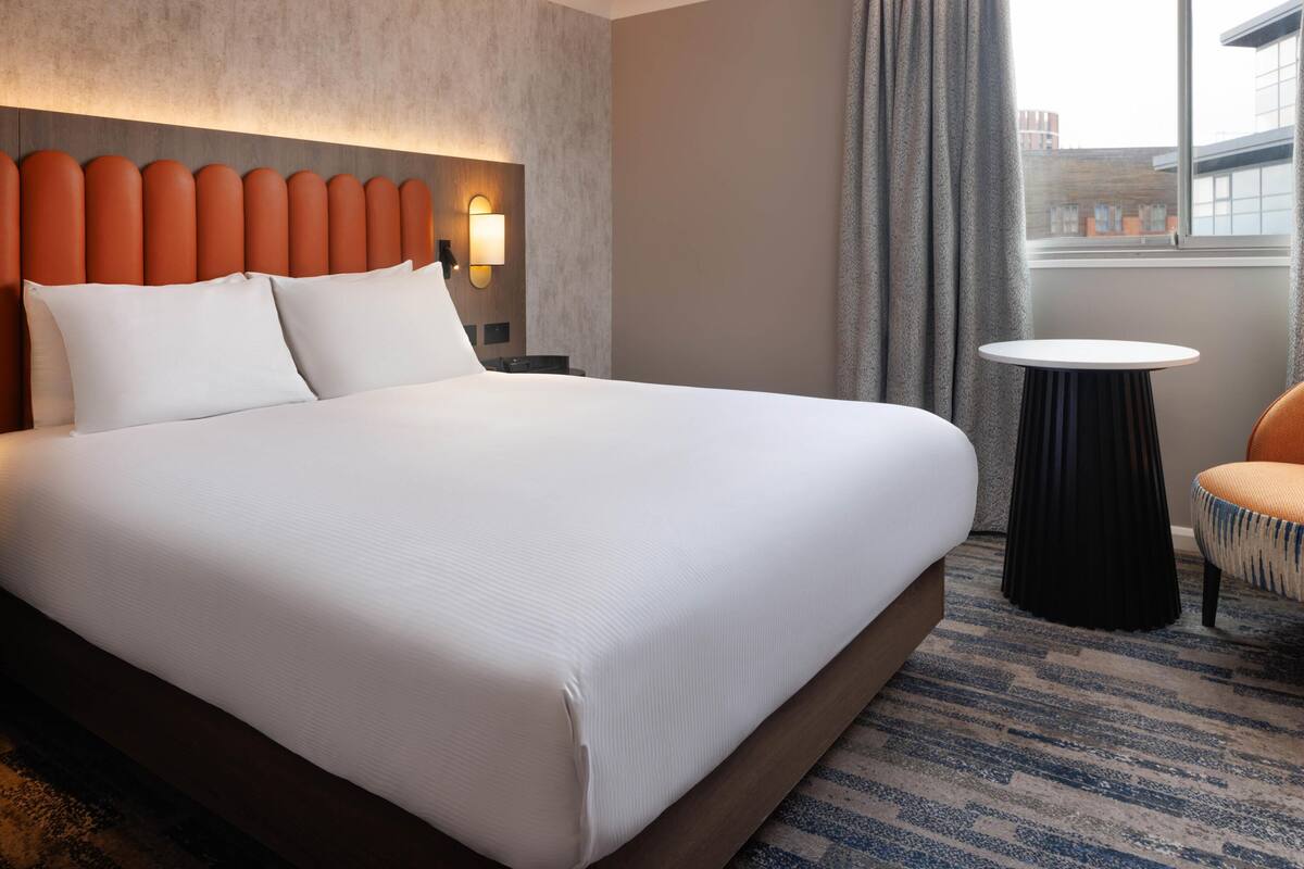 A hotel room with a large bed, a round side table, a chair, a headboard with a light fixture, and a view of buildings through a window.