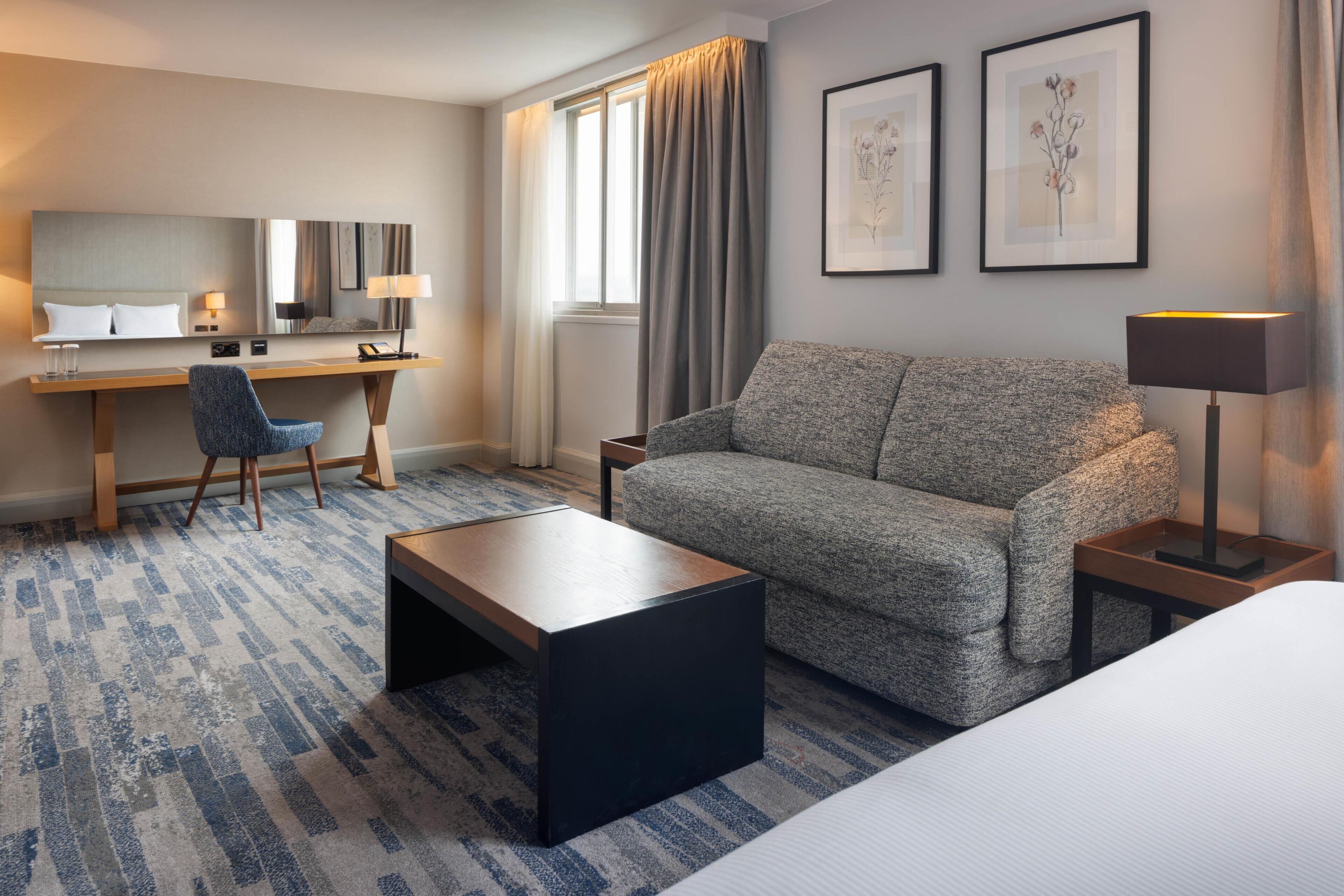 A hotel room with a bed, a sofa, a desk with a chair, a lamp, and two framed pictures on the wall.
