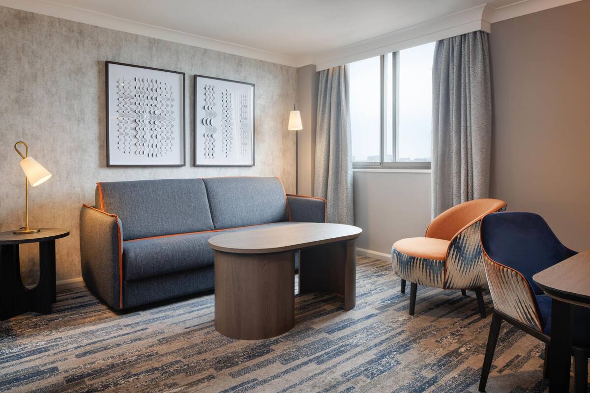 A hotel room with a sofa, armchair, side table, lamp, and artwork on the wall.