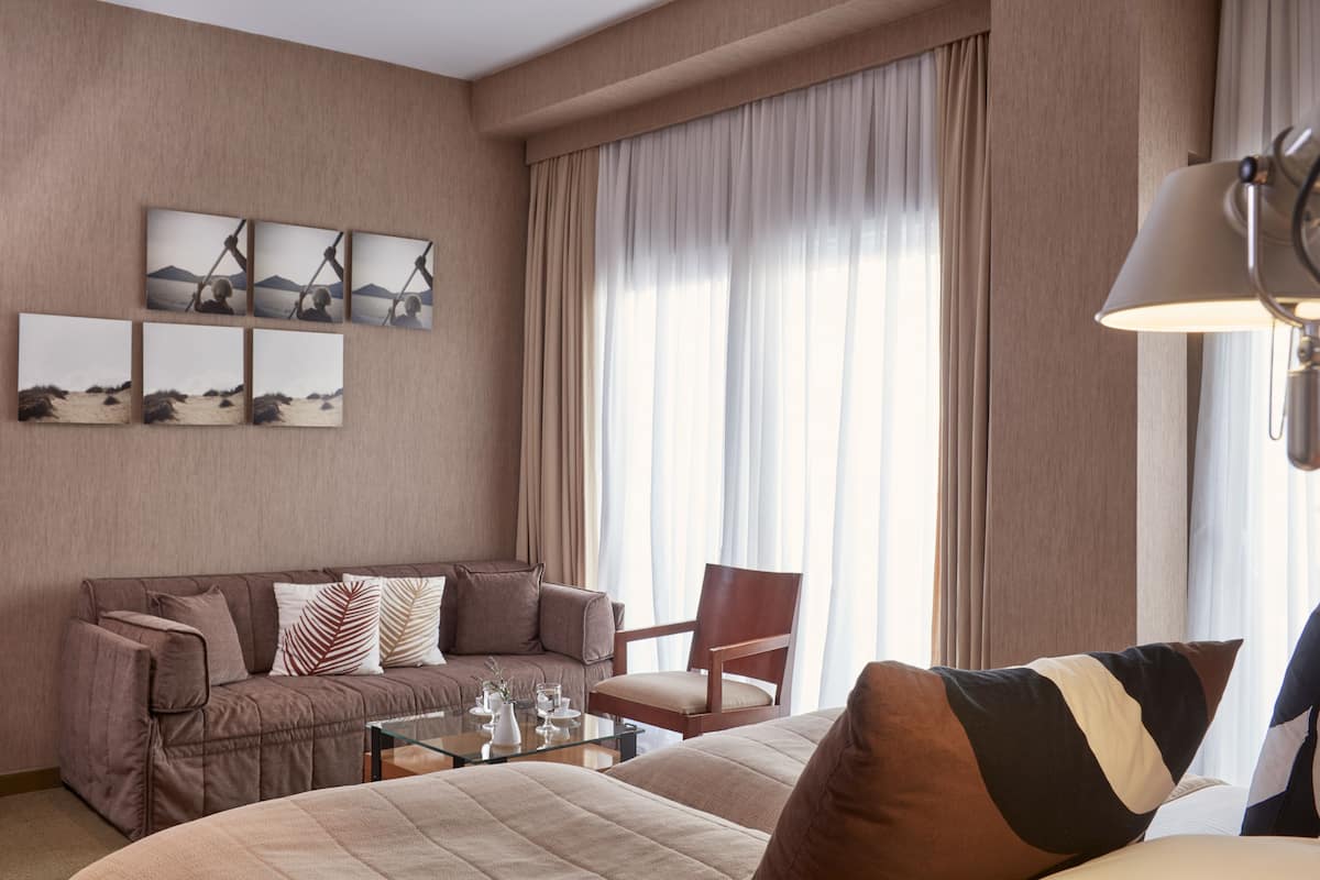 Une chambre d’hôtel avec un lit, un canapé, une chaise, une table basse et une grande fenêtre avec des rideaux.