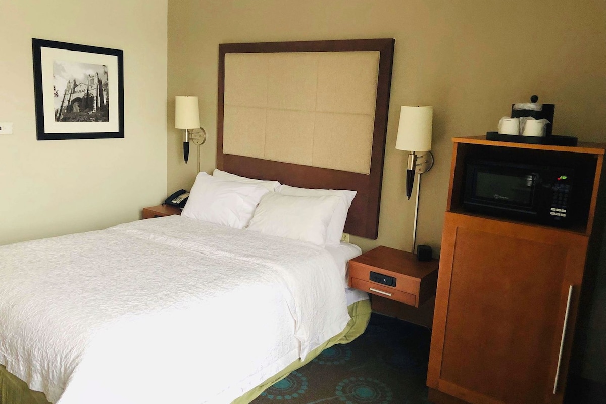 A hotel room with a bed, bedside lamps, a nightstand, a microwave, and a framed picture on the wall.