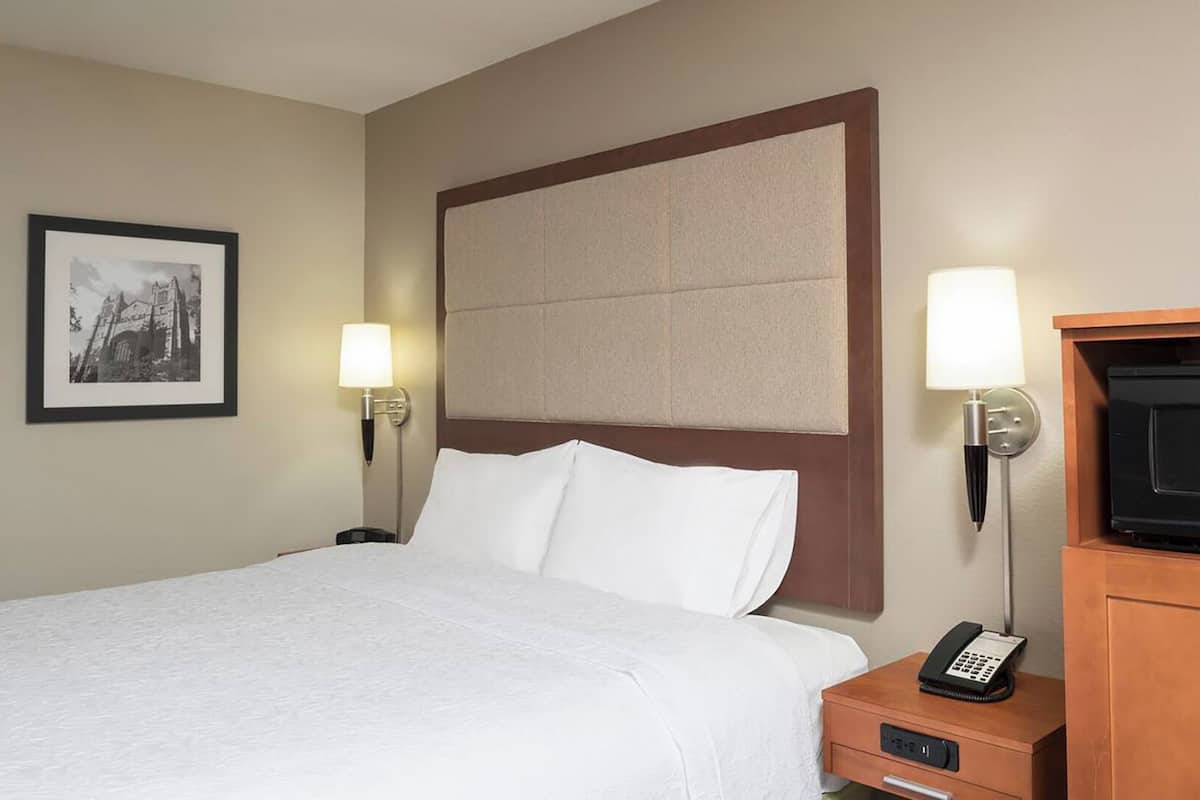 A hotel room with a large bed, two bedside lamps, a framed picture on the wall, a wooden nightstand with a telephone, and a television on a wooden cabinet.
