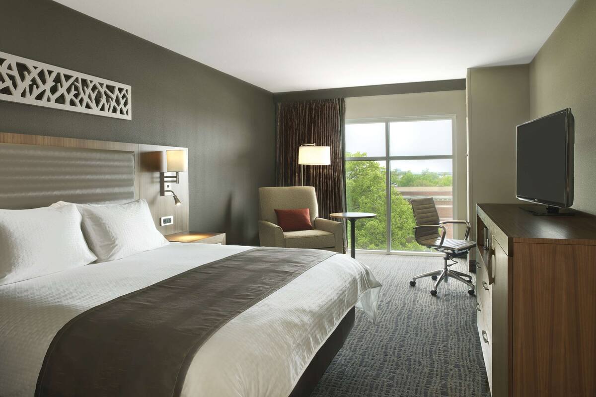 A hotel room with a large bed, a desk with a television, a chair, a small table, and a view of the outdoors.