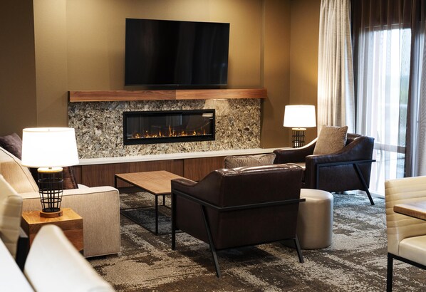 A modern living room with a fireplace, comfortable seating, a flat-screen TV, and a large window with curtains. - The Broadway Columbia - a DoubleTree by Hilton (Columbia)