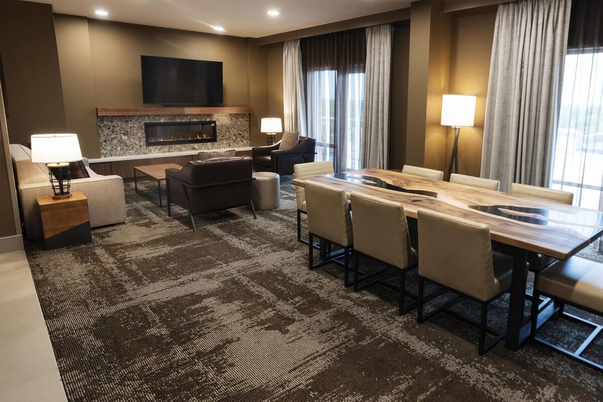 A hotel room with a fireplace, a TV mounted on the wall, a dining area with a wooden table and chairs, and a large window with curtains.