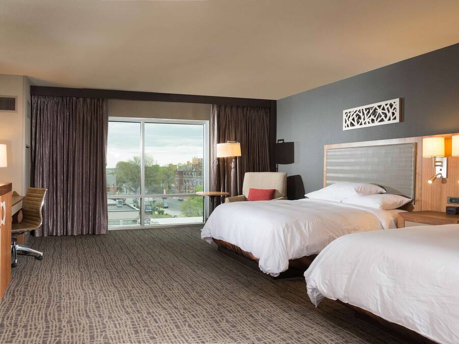 A hotel room with two beds, a TV, a desk, and a view of the city.