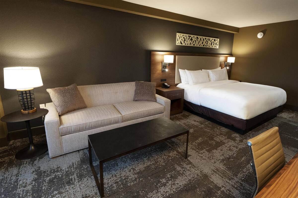 A hotel room with a sofa, a bed, a nightstand with a lamp, and a small table.