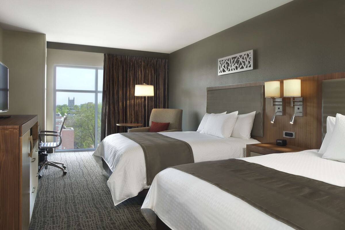 A hotel room with two beds, a desk, a chair, and a view of a building through the window.