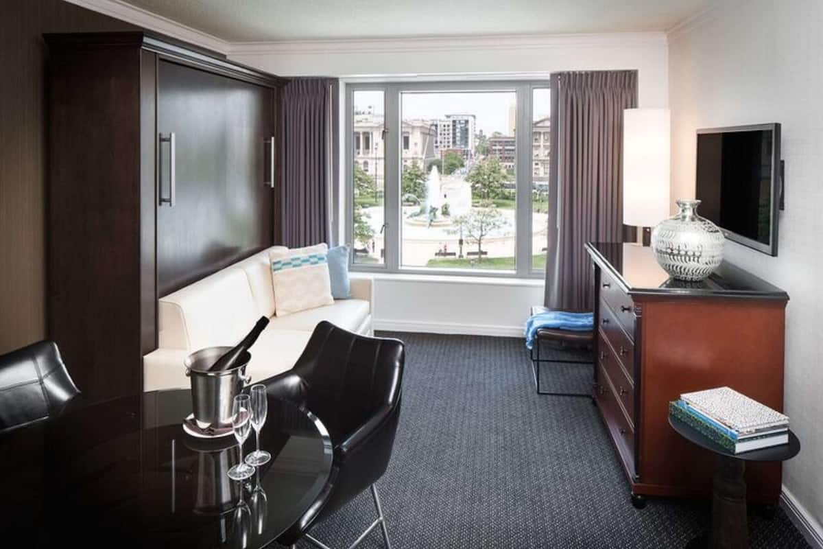 A hotel room with a large window offering a city view, a sofa, a dining table with chairs, a TV, and a minibar.