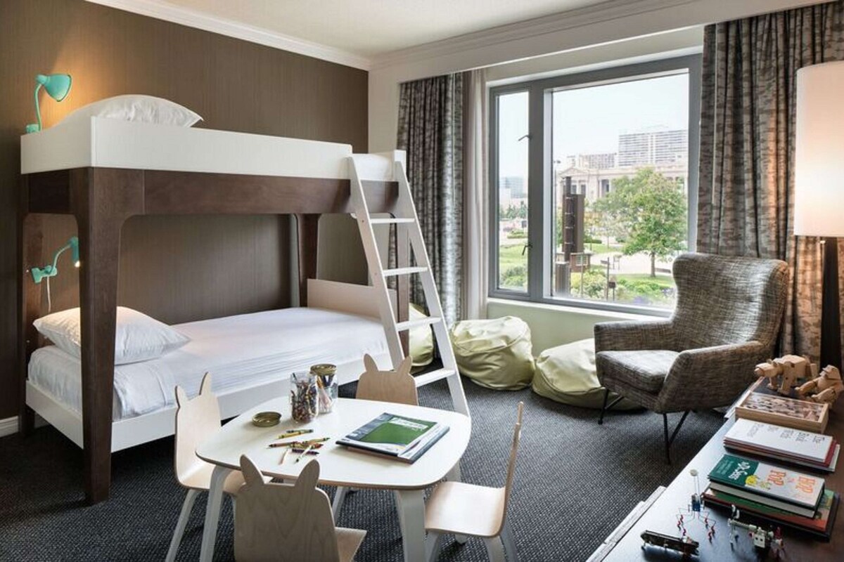 A bunk bed with a ladder, a desk with a chair, a lamp, and a view of the outside through a window.