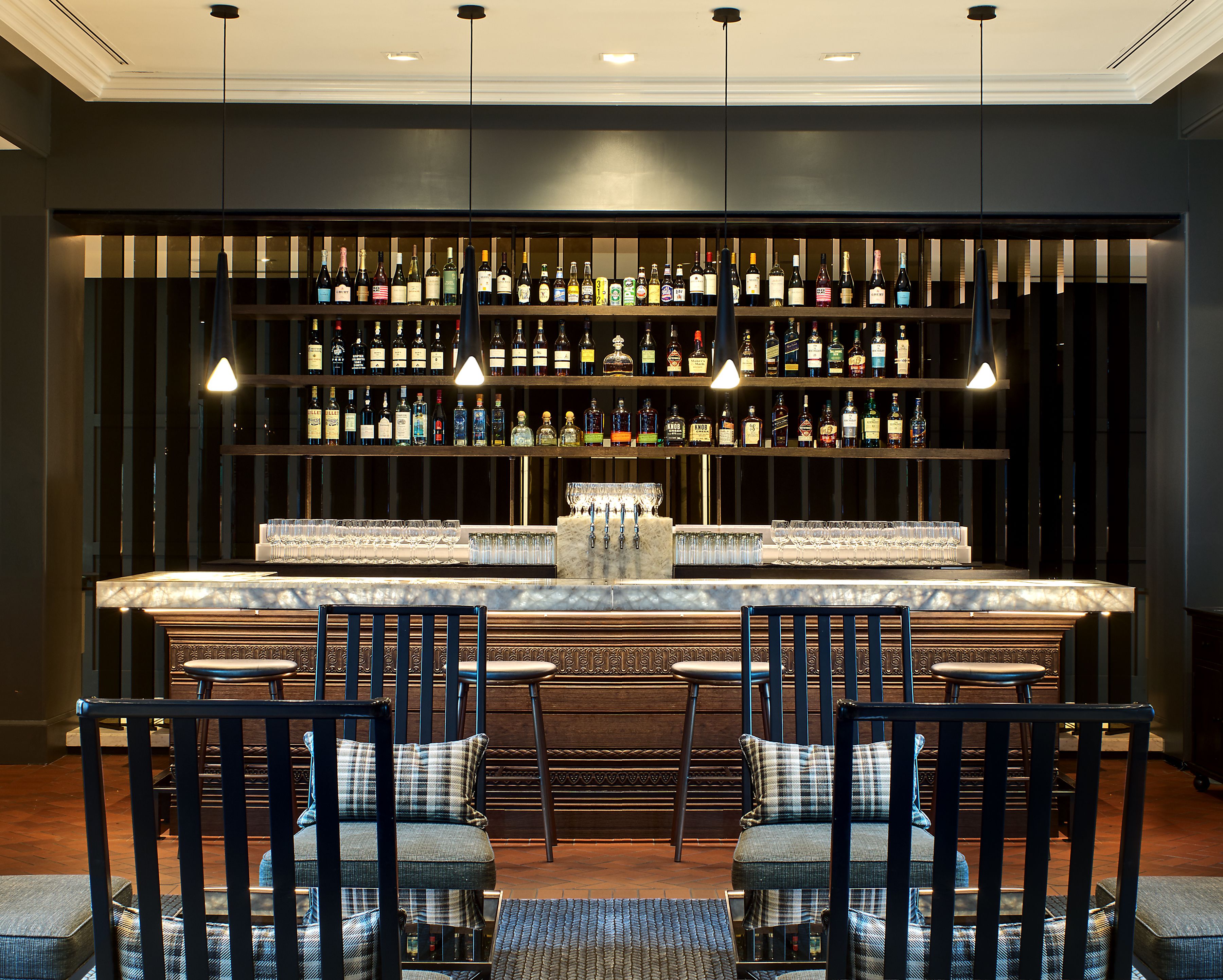 A bar area with multiple shelves of liquor bottles, a marble bar counter, and four upholstered chairs with cushions.