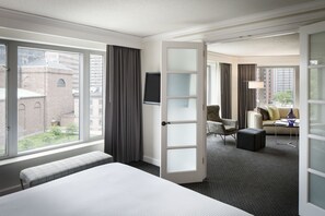 Ein Hotelzimmer mit einem großen Bett, einer Sitzecke inklusive Sofa und Sesseln, einem Fernseher und Blick auf die Stadt durch große Fenster.