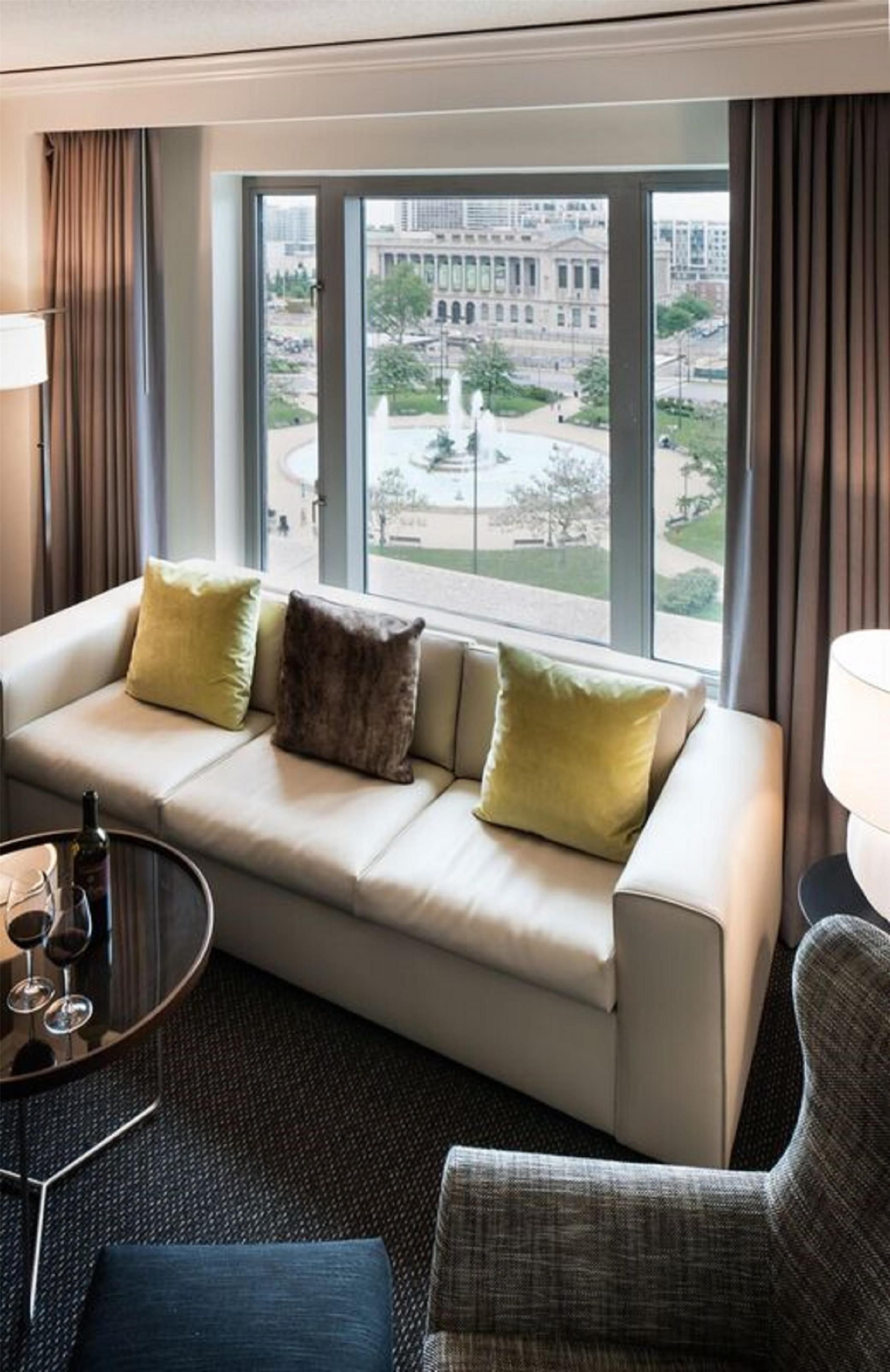 Ein Hotelzimmer mit einer Couch, einem kleinen Tisch mit Flasche und Gläsern sowie Blick auf ein Gebäude und einen Park durch große Fenster.