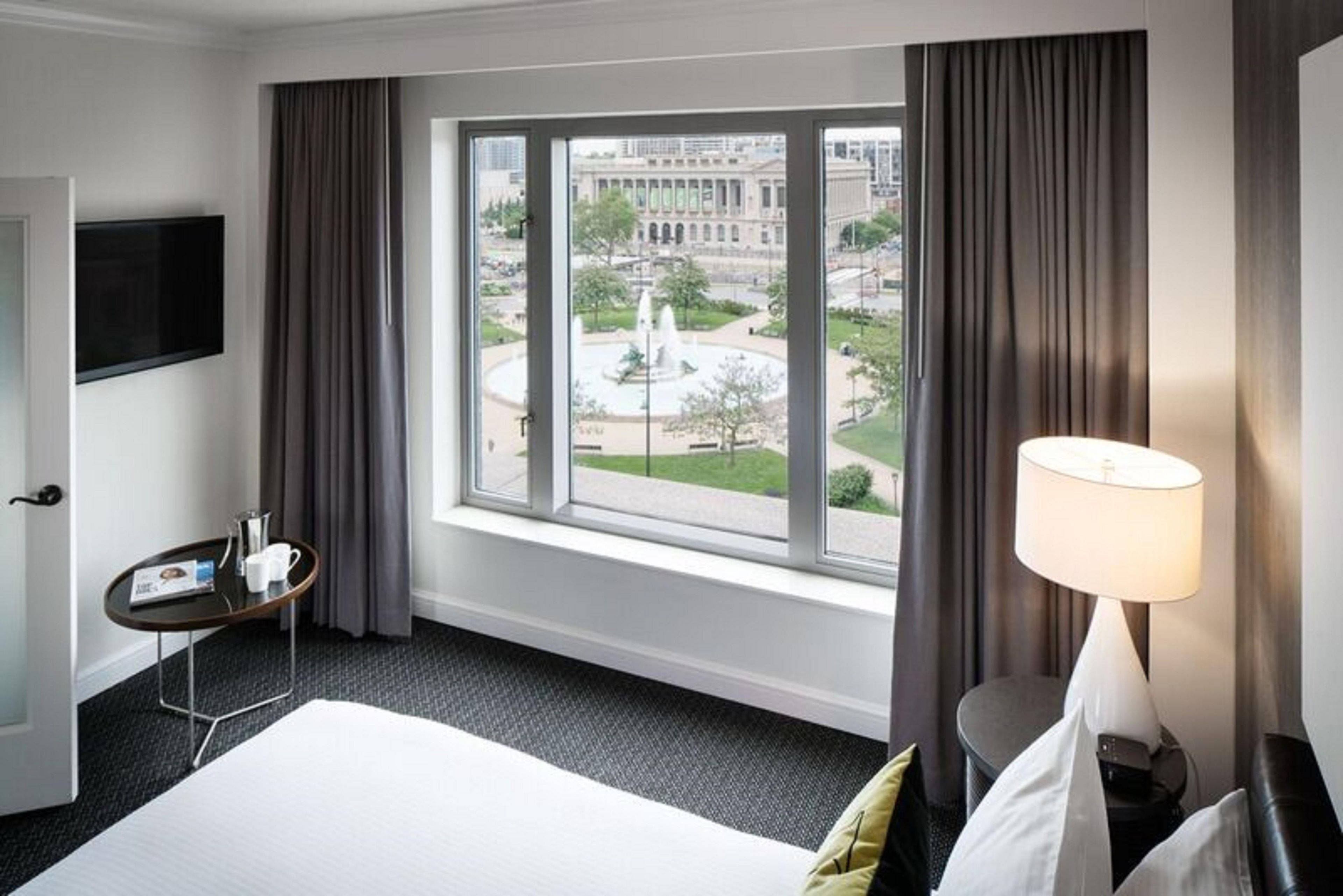 A hotel room with a bed, a small table with a pitcher and cups, a lamp, and a view of a cityscape through a large window.