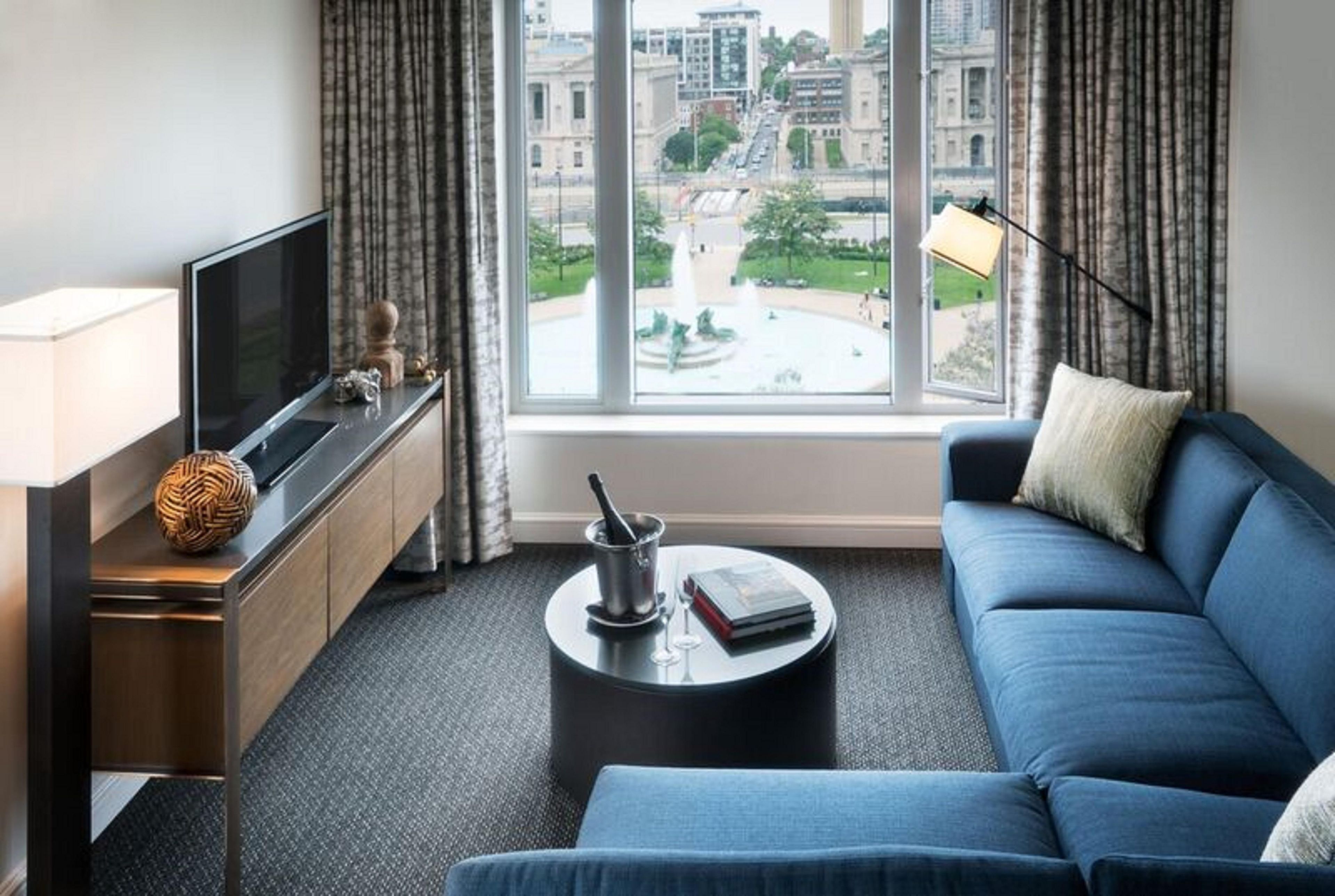 Habitación de hotel moderna con un ventanal grande, televisor de pantalla plana, sofá, mesa de centro y un mueble pequeño.