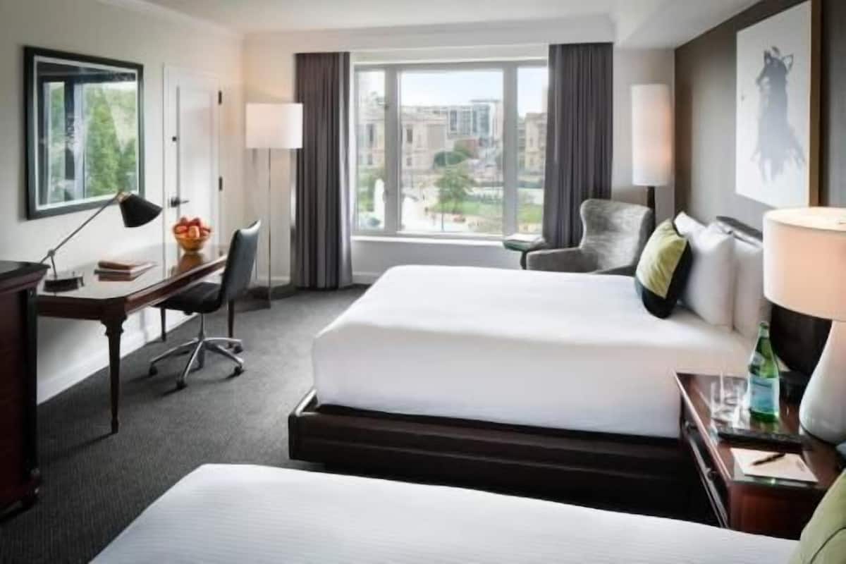 A hotel room with two beds, a desk, a chair, a lamp, and a view of the city.