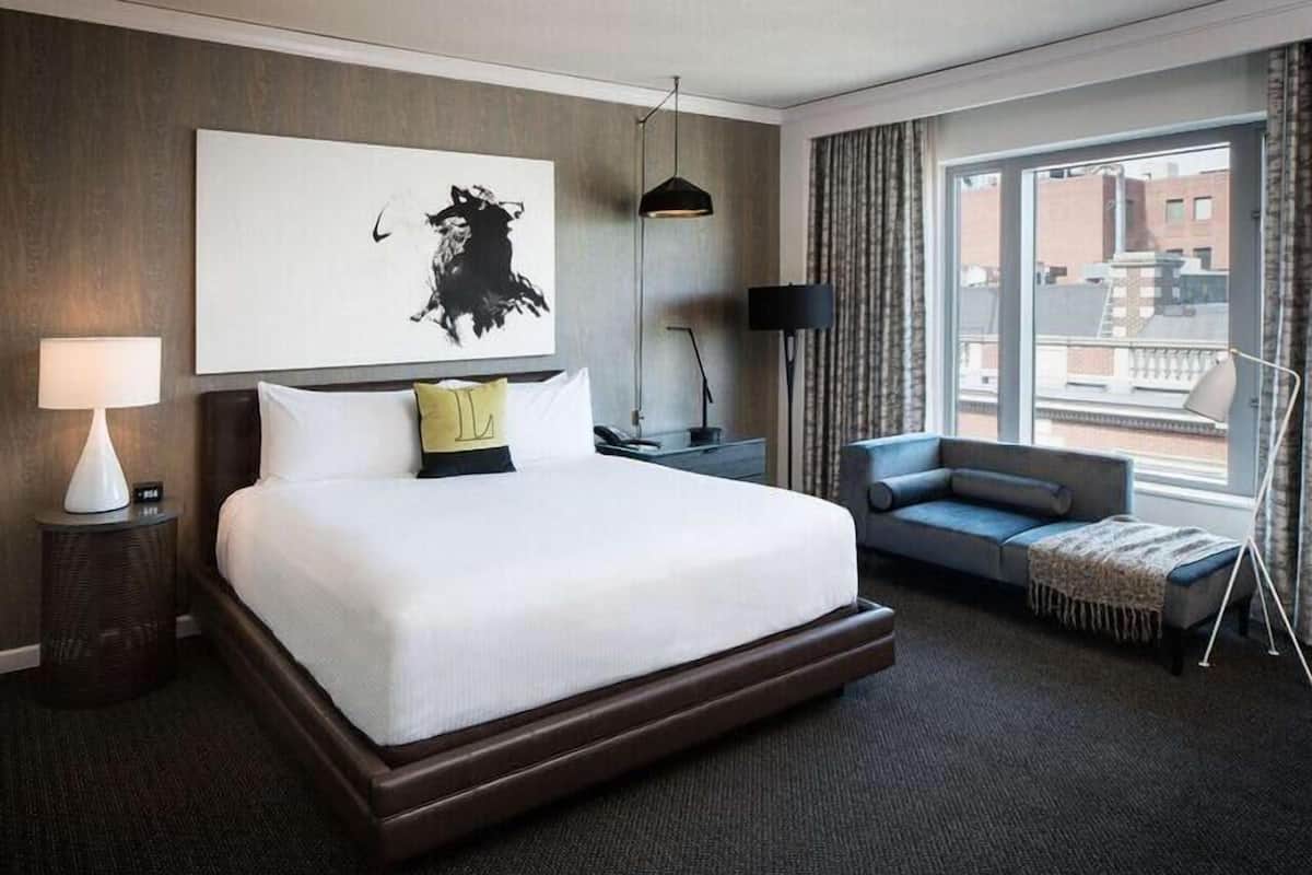 A hotel room with a large bed, a sofa, a desk, a lamp, and a window with a view of buildings.