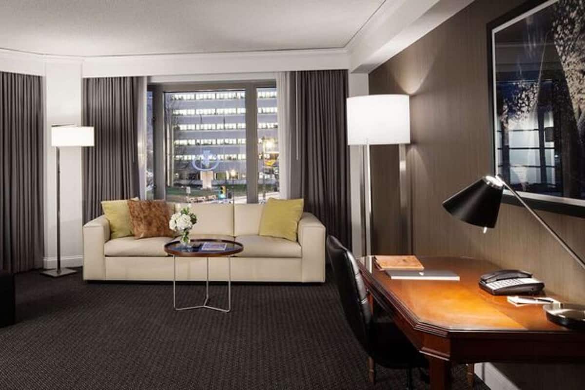 A hotel room with a sofa, armchair, desk, and a view of the cityscape.