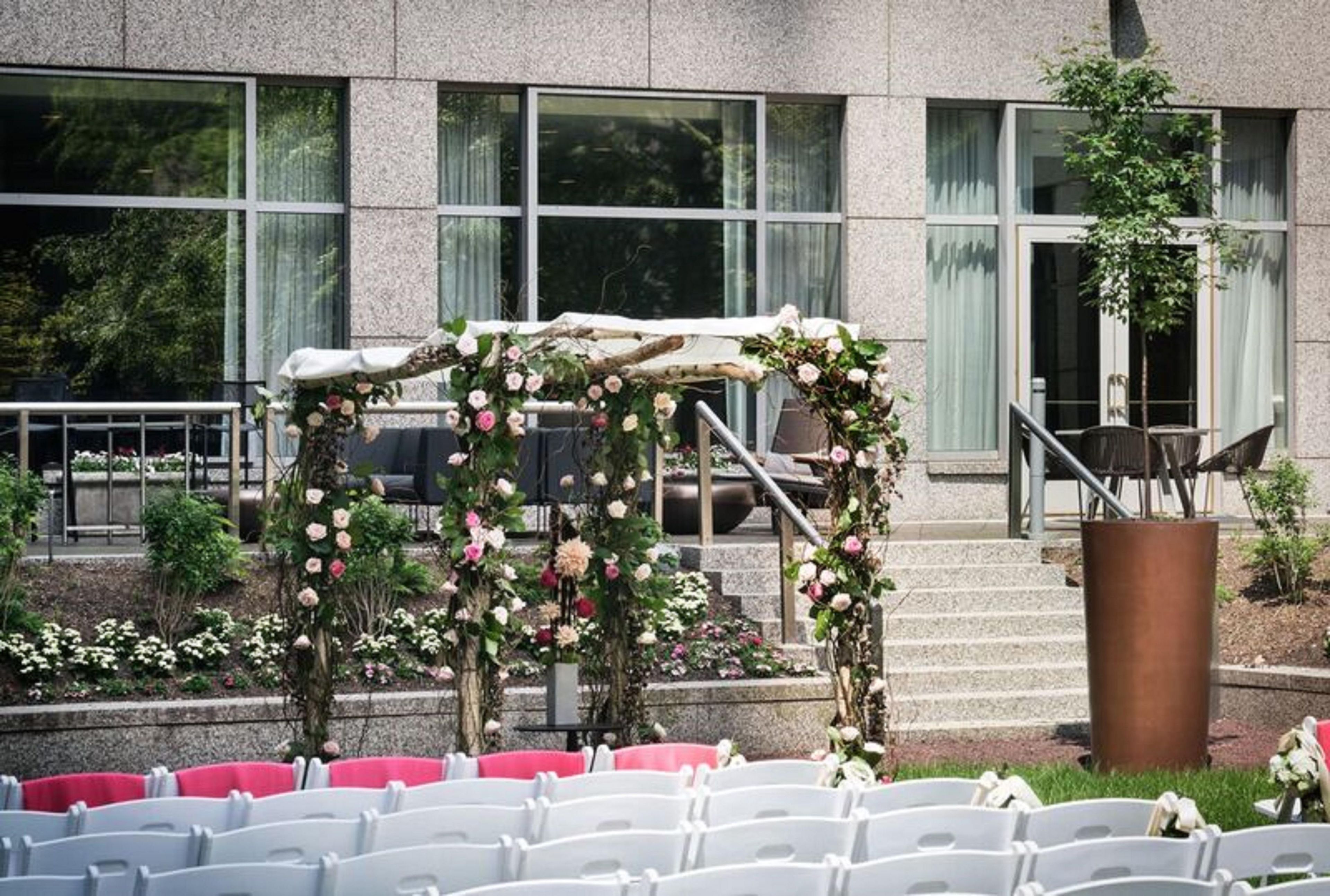 An outdoor seating area with white chairs arranged in rows, a stone building with large windows in the background, and a floral archway.