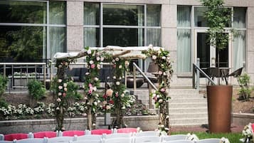 An outdoor seating area with white chairs arranged in rows, a stone building with large windows in the background, and a floral archway.