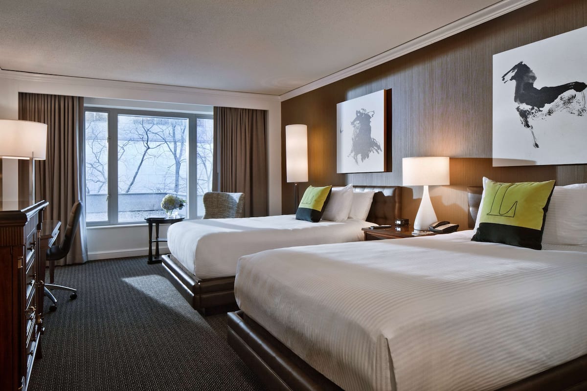 A hotel room with two beds, a desk, a chair, a window with curtains, and a mounted artwork.