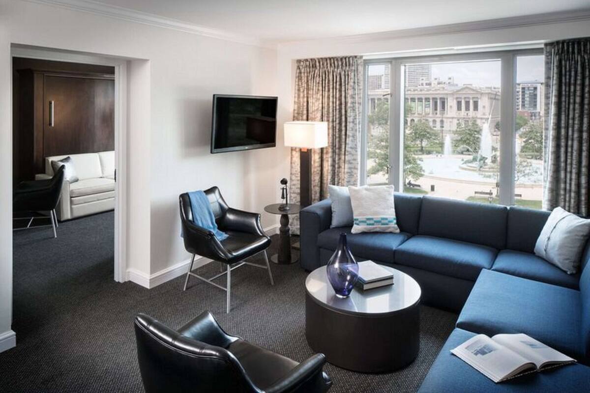 A modern hotel room with a dark blue sofa, a black leather chair, a round coffee table, and a flat-screen TV mounted on the wall.