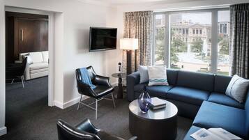 Una habitación de hotel moderna con un sofá azul oscuro, un sillón de cuero negro, una mesa de centro redonda y un televisor de pantalla plana montado en la pared.