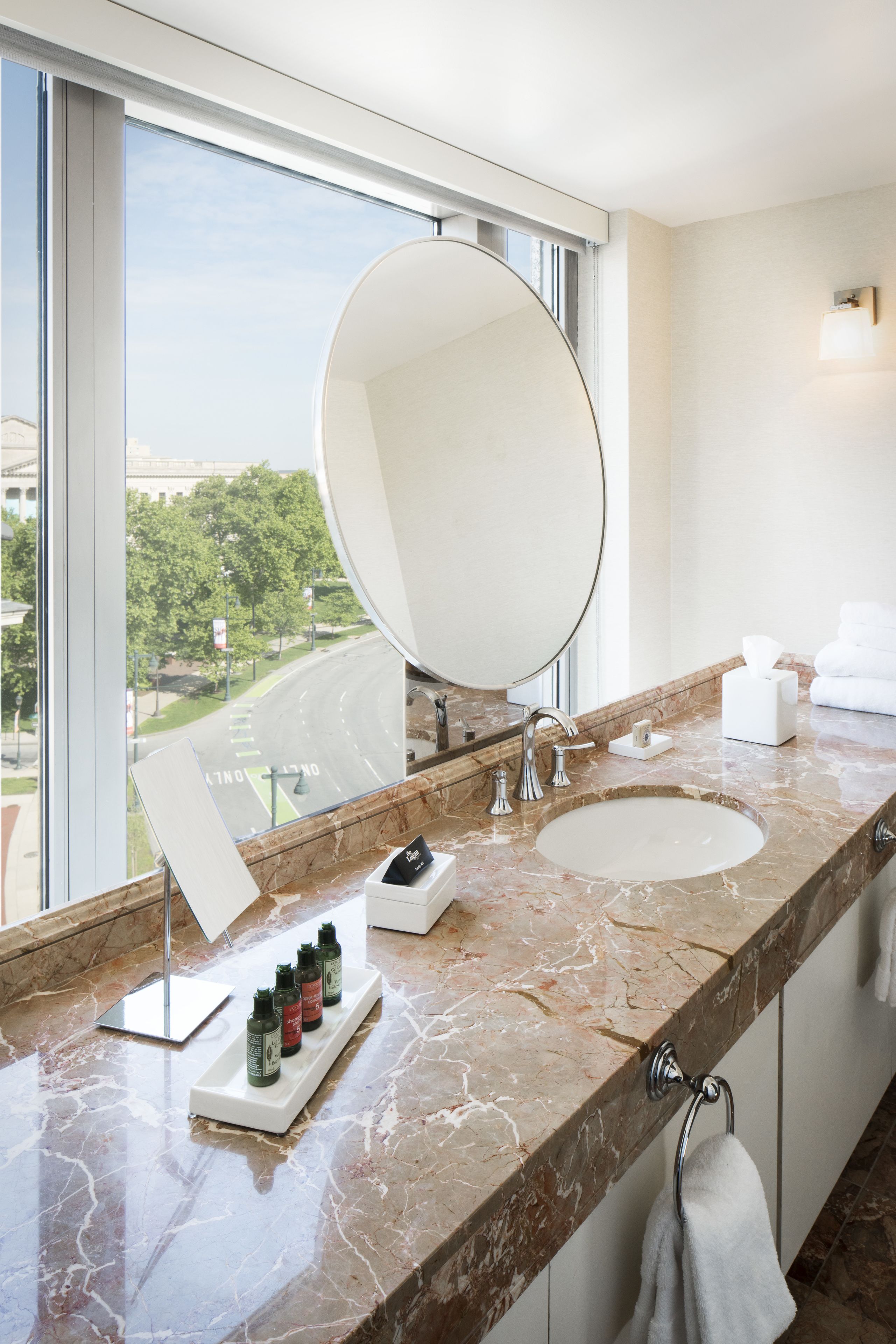 A modern bathroom with a large oval mirror, a sink, and a window offering a view of a road and greenery.