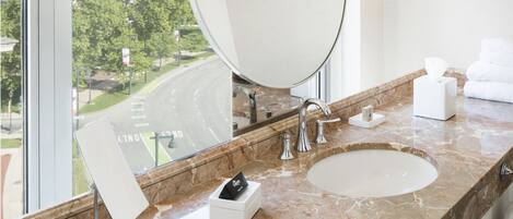 A modern bathroom with a large oval mirror, a sink, and a window offering a view of a road and greenery.
