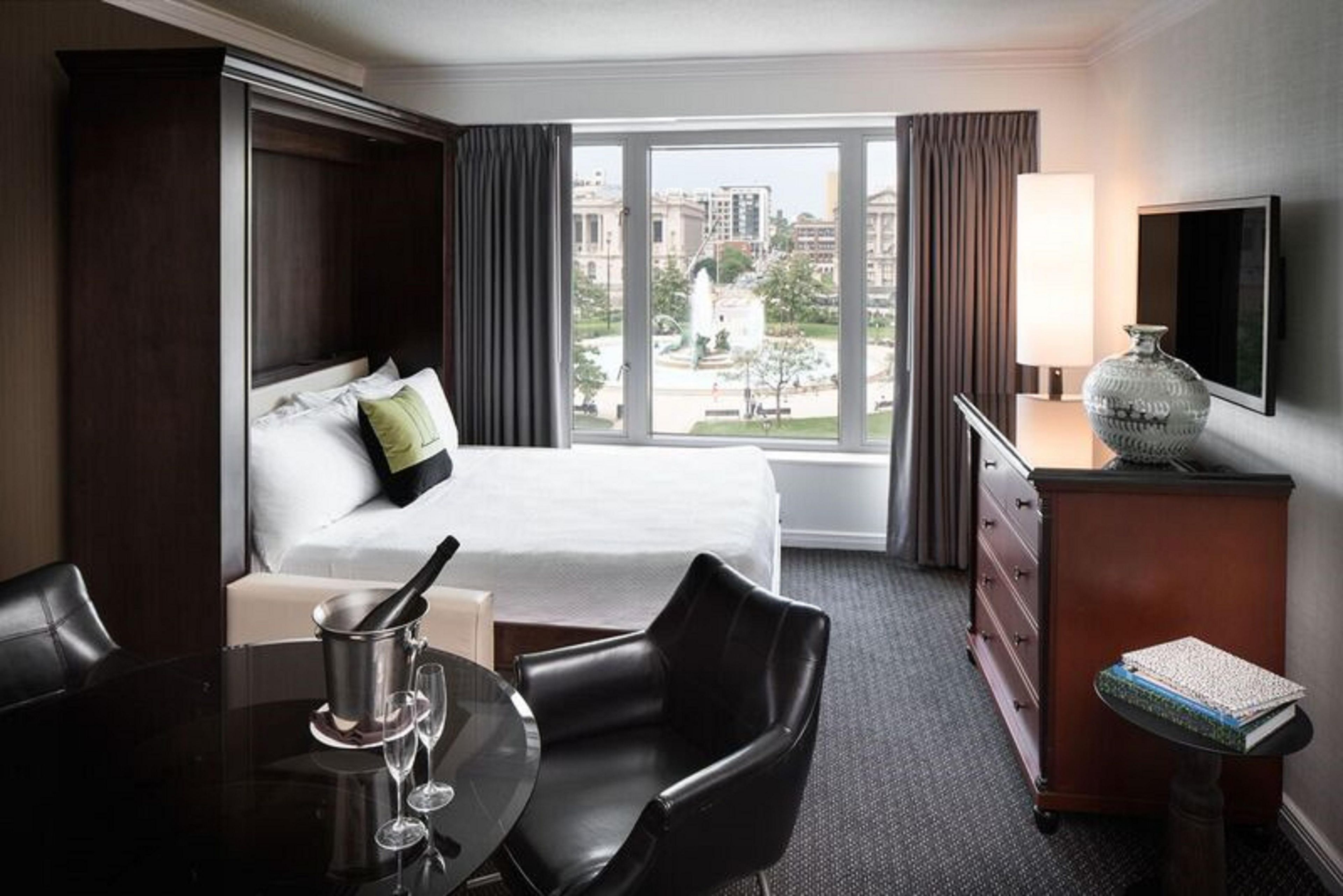 A hotel room with a bed, a desk with a lamp, a chair, a dining table with wine glasses, and a view of the city.