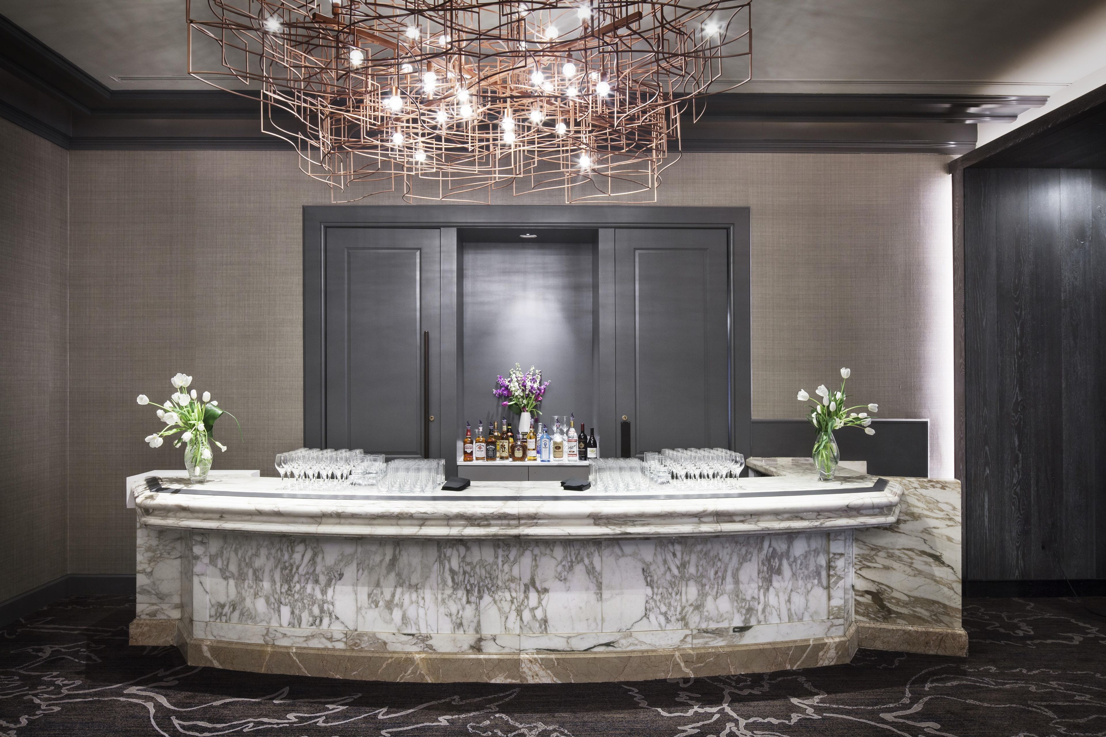 A marble reception desk with a modern chandelier, two vases of flowers, and a display of bottles and glasses.