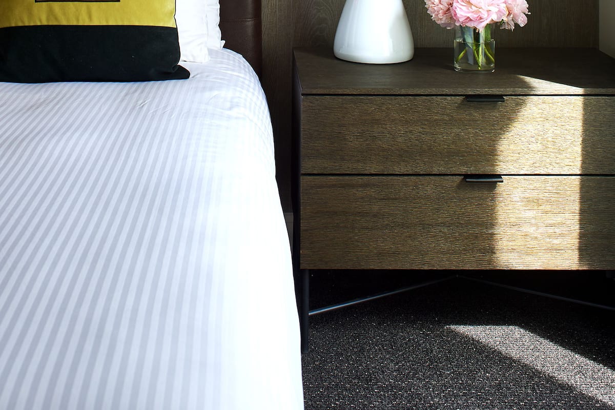 A neatly made bed with striped bedding, a white bedside lamp, a small vase with pink flowers, and a wall art piece above the bed.